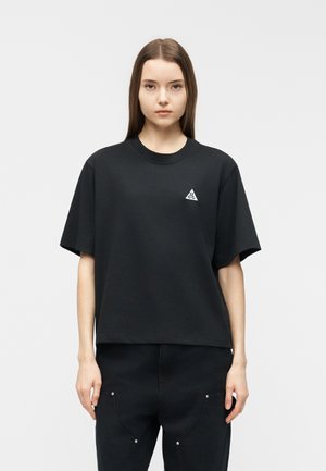 Jeune femme aux cheveux bruns lisses portant un T-shirt noir surdimensionné avec un petit logo triangulaire blanc sur la poitrine.