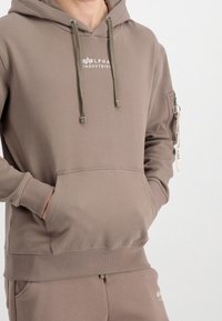 Sweat à capuche couleur sable, en tissu doux. Comprend une poche kangourou, des poignets côtelés et une impression de logo. Poche latérale avec détail fermeture éclair.