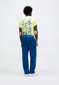 Κίτρινη γραφική μπλούζα με εικονογραφήσεις στην πλάτη, συνδυασμένη με μπλε φόρμες. Διαθέτει χαλαρή εφαρμογή και ζωνάκι με κορδόνι στη μέση.