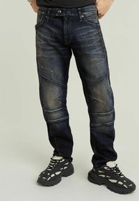 Dark blue denim jeans with a tapered fit, featuring angled seam details and a faded texture. Worn with black sporty shoes.