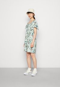 Vestido com padrão floral em tecido verde-claro, mangas curtas e decote quadrado, combinado com ténis brancos de cano alto e um chapéu de balde bege.