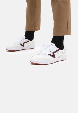 White sneakers with a maroon accent, smooth leather, perforated toe box, and rubber sole. Styled with brown pants and black socks.