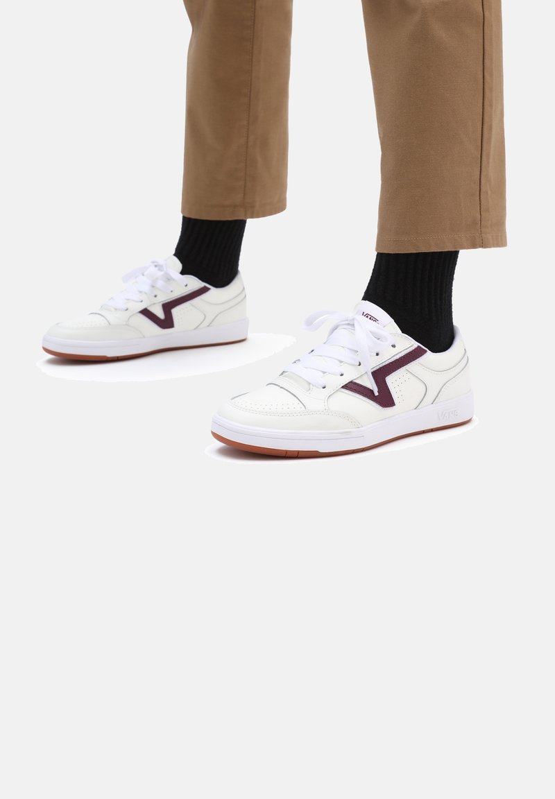 Baskets blanches avec un accent bordeaux, cuir lisse, embout perforé et semelle en caoutchouc. Stylées avec un pantalon marron et des chaussettes noires.