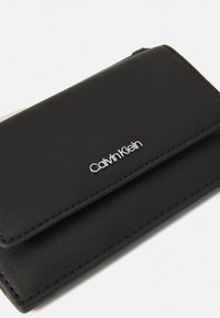 Portefeuille en cuir noir avec une surface texturée, une fermeture magnétique et un logo Calvin Klein argenté à l'avant.