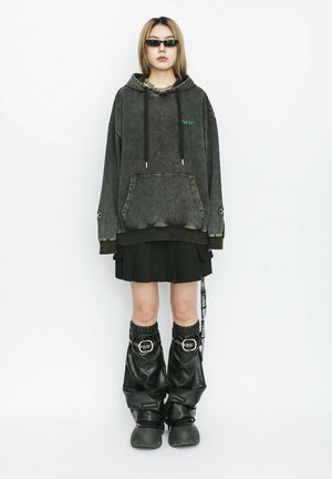 Sudadera gris de gran tamaño con bolsillo frontal, superpuesta a una falda negra con pliegues. Medias brillantes negras y calzado negro robusto completan el look.
