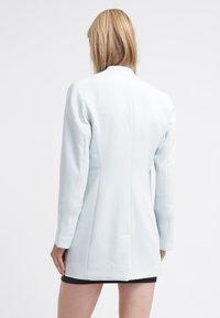 Žena s blond vlasy, oblečená v světlomodrém upraveném dlouhém saku a krátké černé sukni, stojící zády k bílé pozadí.