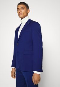 Maßgeschneiderter Herrenanzug aus tiefblauem Wollstoff, mit einem Knopf, spitzen Revers und vorderen Taschen. Getragen über einem weißen Rollkragenpullover.