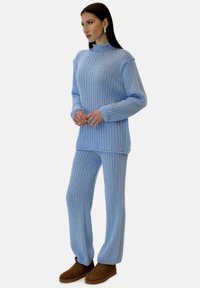 Jasnoniebieski sweter z prążkowanej dzianiny i pasujące spodnie, z wysokim dekoltem i długimi rękawami, uzupełnione brązowym obuwiem. Miękka, teksturowana tkanina.