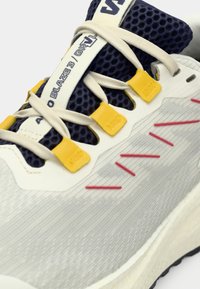 Urheilukenkä, jossa on harmaa verkko-osa, tummansinisiä yksityiskohtia, keltaiset nauhalukot, punaiset ompeleet ja teksturoitu valkoinen pohja mustalla pidolla.