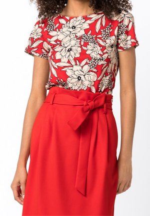 Femme portant un haut à manches courtes rouge à fleurs, rentré dans une jupe taille haute rouge avec une ceinture en tissu nouée.