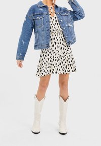 Denimjack met versleten accenten, witte jurk met een zwart bladpatroon en kniehoge witte laarzen met een glanzende afwerking.