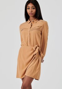 Robe en bronze à manches longues, avec des détails plissés et une taille nouée. Fabriquée en tissu léger avec une texture lisse.