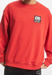 Rode sweatshirt met lange mouwen, ribgebreide manchetten en zoom, met een zwarte rechthoekige logopatch op de linkerborst. Zachte katoenmix.