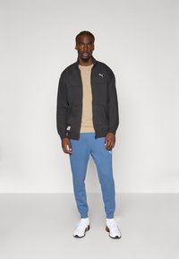 Jachetă neagră cu fermoar peste o cămașă bej, combinată cu pantaloni de trening albastru deschis. Tenisi albi cu talpă neagră. Design simplu, athletic.