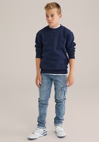 Tumši zils sporta krekls ar ribotām aprocēm, kombinēts ar gaiši zilu džinsiem ar sānu kabatām, un baltām sporta apaviem ar ziliem akcentiem.