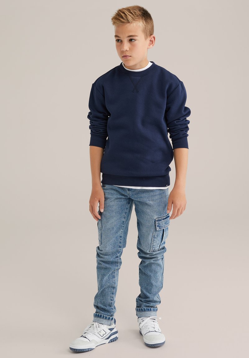 Tumši zils sporta krekls ar ribotām aprocēm, kombinēts ar gaiši zilu džinsiem ar sānu kabatām, un baltām sporta apaviem ar ziliem akcentiem.