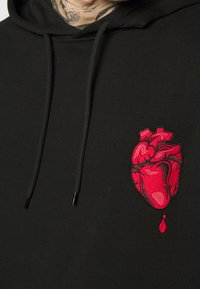 Sudadera negra con un corazón anatómico rojo bordado en el pecho, goteando una sola gota de sangre por debajo.