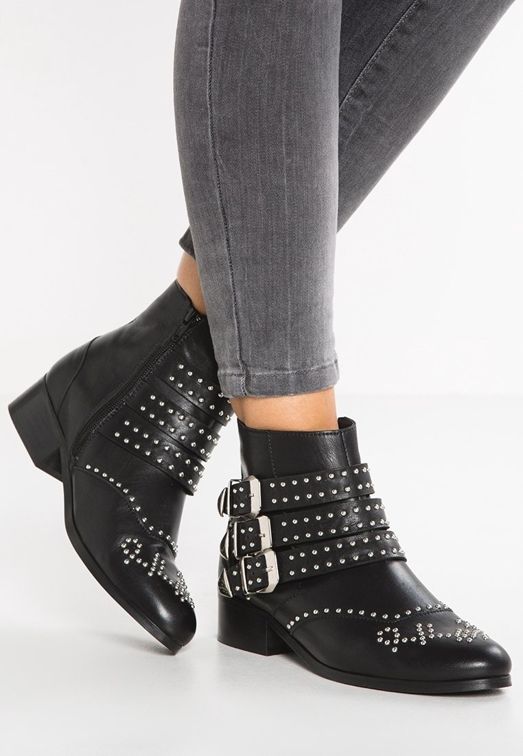 Bottines noires à la cheville avec trois brides bouclées et des clous argentés portées avec un jean skinny gris sur fond blanc.
