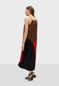 Abito maxi plissettato con blocchi di colore marrone, rosso e nero. Spalline regolabili e un nastro sul retro. Indossato con sandali neri con borchie.