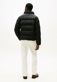 Giacca a vento nera, con design trapuntato, collarino alto e maniche lunghe. Abbinata a jeans bianchi e scarpe scure. Sfondo neutro.