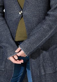 Henkilö, joka pitää molemmilla käsillä tummansinistä ja valkoista teksturoitua neuleliiviä, jonka edessä on yksi nappi, sekä oliivinvihreää paitaa ja sinisiä farkkuja.