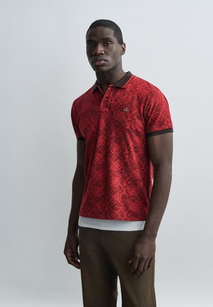 Homme portant un polo rouge à motifs avec un col et des bas de manches noirs, associé à un pantalon marron, debout devant un fond clair uni.
