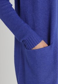 Blå stickad cardigan med ribbade muddar och en framficka, med en mjuk textur och en avslappnad passform.