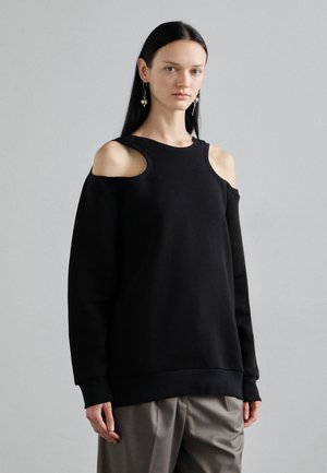 Black sweatshirt with cut-out shoulder design, ribbed neckline, and cuffs. Fabric appears soft and textured, paired with loose, beige trousers.