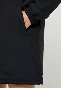 Handen i fickan på en svart jacka med elastisk mudd och liten Vans-logotagg nära fållen på en ljus bakgrund.