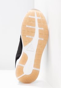 Semelle de basket noire montrant la bande de roulement beige avec des sections en caoutchouc blanc et un logo triangulaire au centre, positionnée contre des murs blancs d'angle.