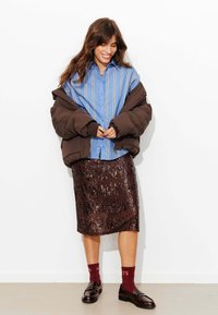 Doudoune marron, chemise boutonnée à rayures bleues, jupe à sequins, chaussettes rouges avec des chiffres et mocassins noirs sur fond blanc.