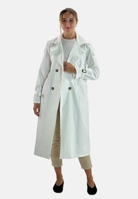 Witte trenchcoat met een dubbelgeknoopt ontwerp, grote kraag, knoopdetails en manchetriemen, gedragen over een licht topje en beige broek.