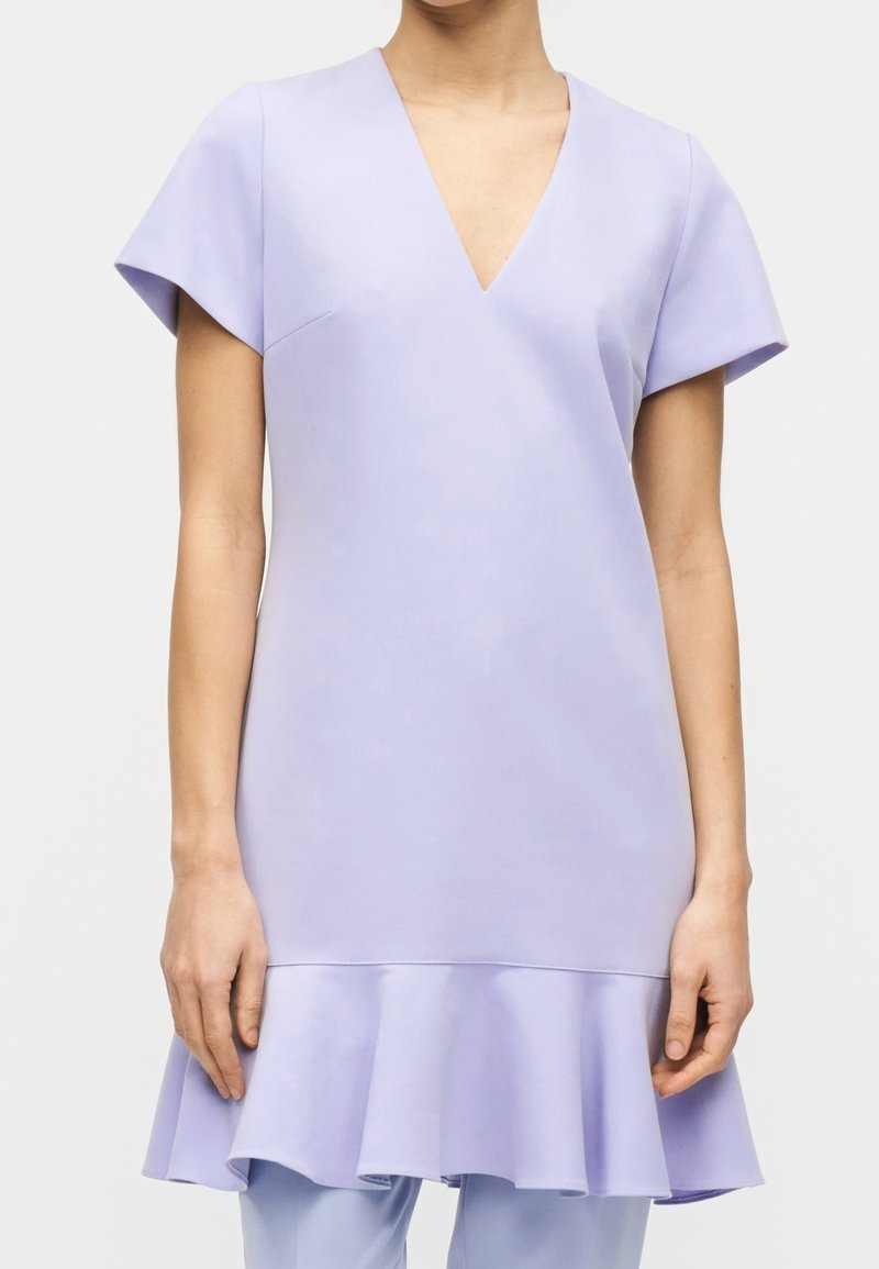 Femme portant une robe courte à manches courtes violet clair avec un col en V et un ourlet évasé, debout devant un fond uni.