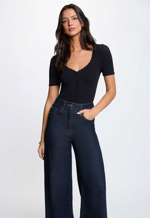 Kvinde med langt mørkt hår iført en sort kortærmet knapbluse og højtaljede vidde mørkeblå jeans, den ene hånd i lommen.