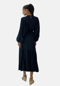 Schwarzes langärmliges Kleid mit gerafftem Oberteil, gestuften Rock und Schlüsselrücken-Detail, kombiniert mit tanfarbenen Keilabsätzen. Glatte Stoffoberfläche.