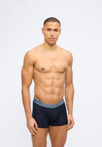 Άνδρας μοντέλο με κοντά μαλλιά που φοράει μαύρα μπόξερ Tommy Hilfiger, στέκεται μπροστά σε μια απλή ανοιχτόγρια φόντο.