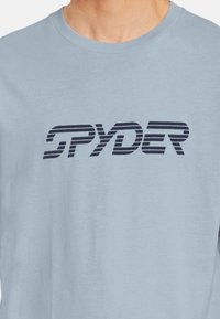 Camiseta de manga larga azul claro, hecha de algodón suave, con un logo negro de "SPYDER" a rayas en el pecho. Diseño simple y casual.