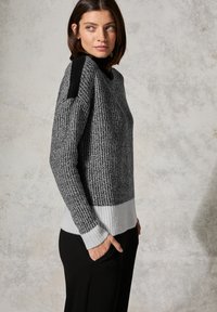 Pull en tricot noir, gris et blanc avec ourlet et poignets côtelés, présentant un motif texturé et un accent au niveau des épaules. Associé à un pantalon noir.