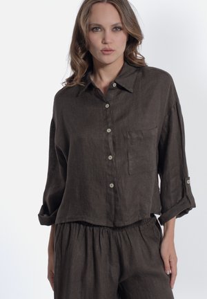 Femme aux cheveux ondulés porte une chemise ample brun foncé à boutons avec poche et un pantalon assorti à taille élastique sur un fond uni.