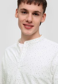 Bílá košile na knoflíky s lehkým materiálem; má malé zelené vzory, stojací límec a hladkou texturu.