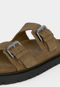 Καφέ σουέντ σανδάλι με δύο ρυθμιζόμενες ασημένιες αγκράφες και χοντρή μαύρη σόλα, με τη φίρμα "CPH STUDIOS" στο πλάι.