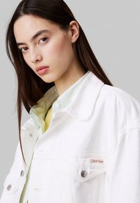 Chaqueta de denim blanca con costuras visibles y botones a presión. Presenta un bolsillo frontal, etiqueta con el logo, y se lleva con una camiseta verde clara debajo.