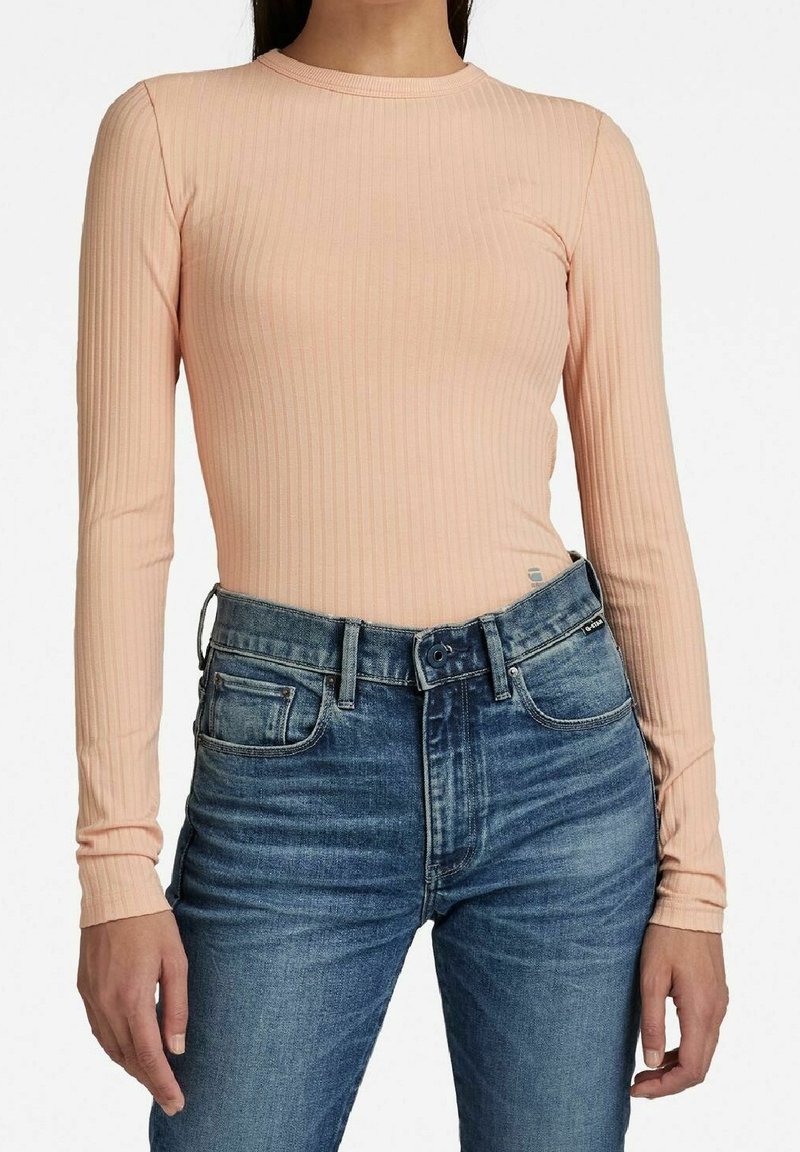 Długi rękaw top prążkowany w miękkim brzoskwiniowym kolorze. Dopasowany fason z okrągłym dekoltem, zestawiony z wysokim stanem niebieskich jeansów.