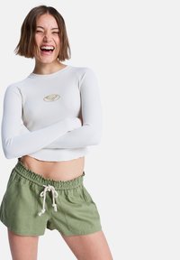 Haut court à manches longues blanc côtelé avec un logo doré, associé à un short vert extensible avec une taille froncée et un cordon de serrage.