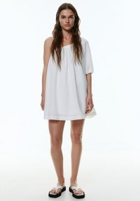 Robe blanche à une épaule avec un tissu texturé, coupe ample et fronces à la taille. Assortie à des tongs blanches avec une semelle noire.