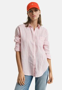 Camicia a righe rosa e bianche con bottoni, realizzata in un tessuto leggero, dotata di taschino e maniche arrotolate, abbinata a un berretto arancione brillante.