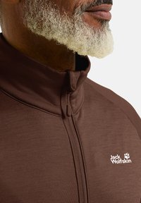 Braune Zip-Jacke mit hohem Kragen, aus glattem Stoff gefertigt. Verfügt über ein kleines weißes Logo auf der Brust und eine subtile Textur.