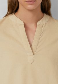 Beige, kragenloses Top aus weichem Stoff mit einem V-Ausschnitt, subtiler gerippter Textur und sichtbaren Nähten entlang der Kanten.