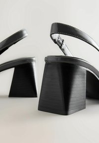 Sandales à talons hauts en cuir verni noir avec talons blocs et brides à la cheville sur un fond gris clair.
