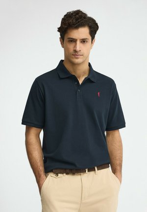 Uomo con capelli ricci scuri che indossa una polo blu navy con un logo rosso e pantaloni beige, in piedi con le mani nelle tasche.
